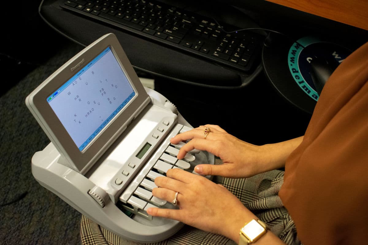 Hands typing on a gray stenotype machine displaying shorthand text on its small screen.